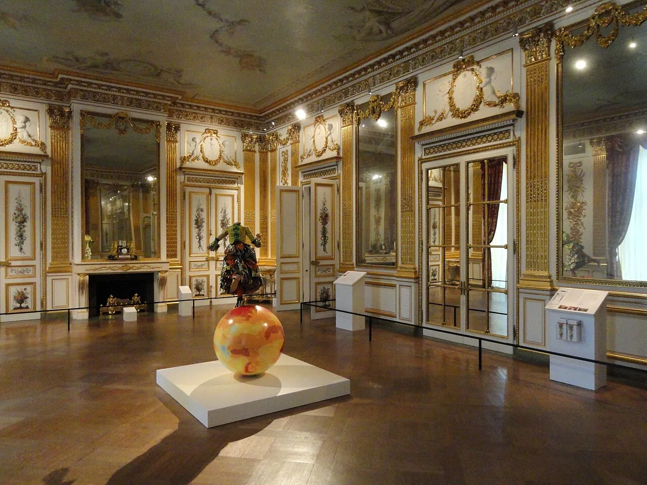 An ornate gallery interior with artworks, interpretive panels, barriers, and a central sculpture