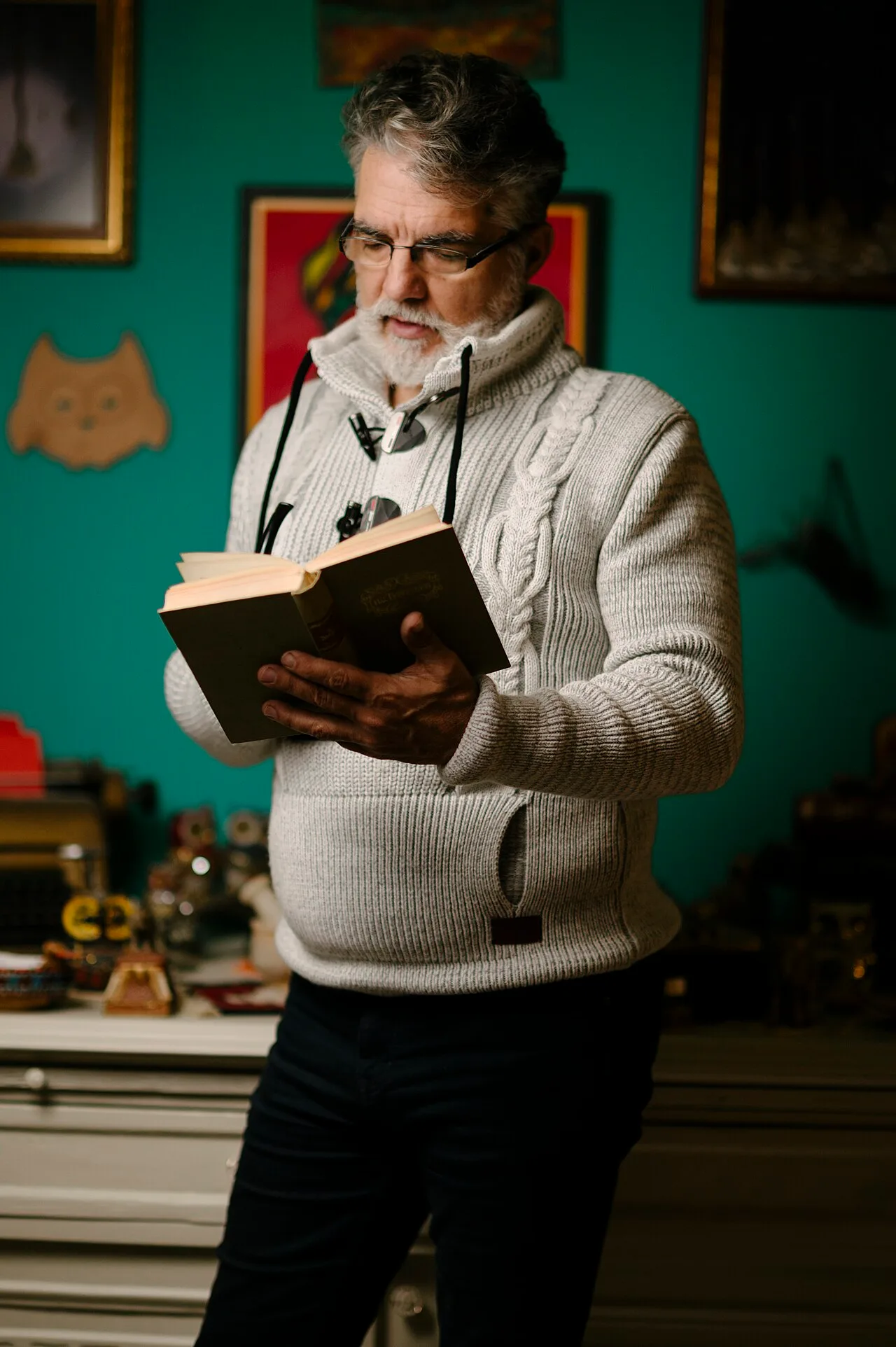 A man in glasses and a knit sweater standing indoors while reading an open book